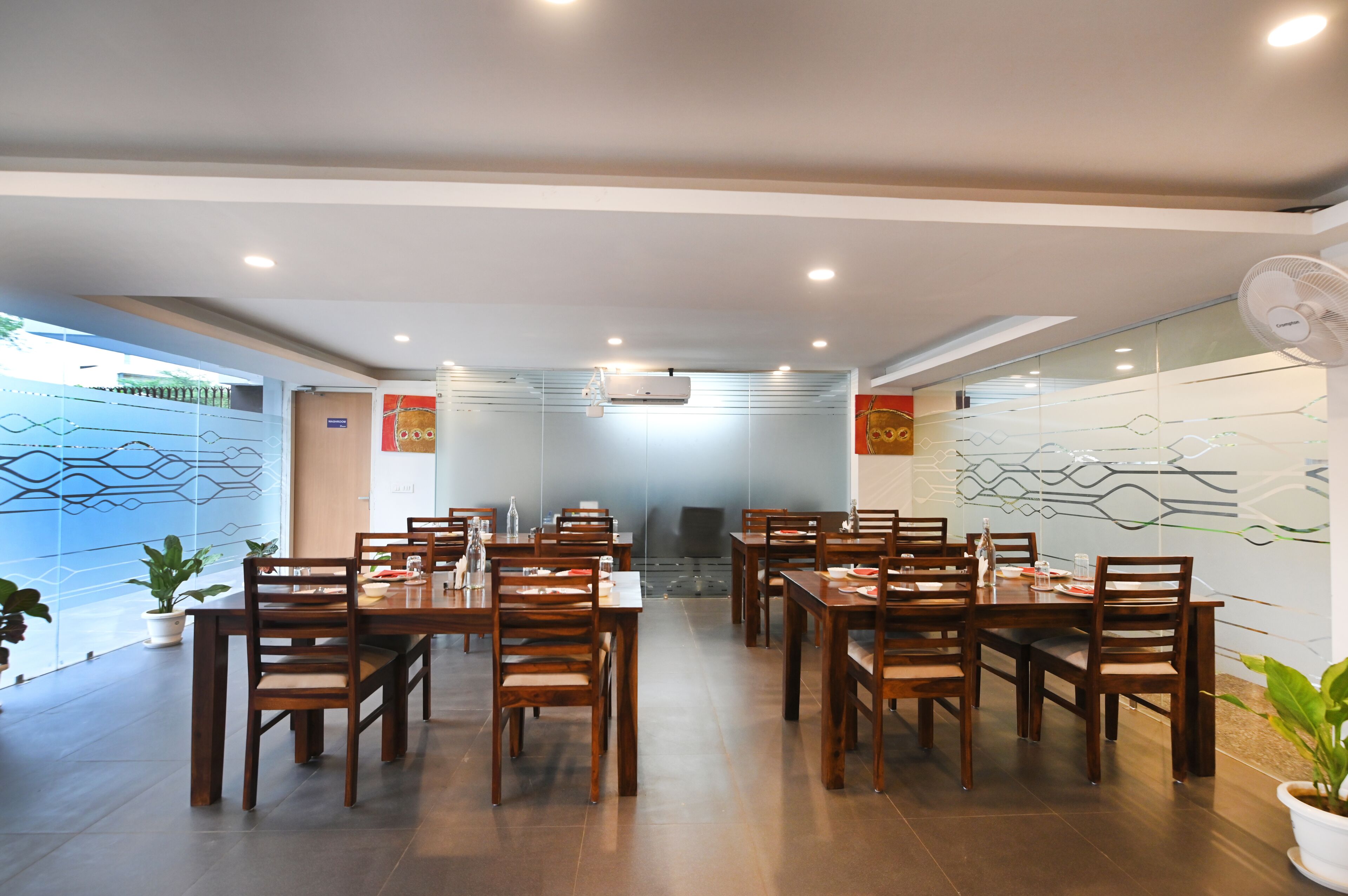 A modern dining area with wooden tables and chairs, glass partitions, and ceiling fans.