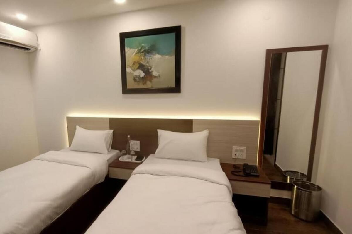 A hotel room with two beds, a painting on the wall, a bedside table with a lamp, a mirror, and an air conditioning unit.