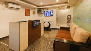 A reception area with a wooden reception desk labeled "The Altruist Business Stays," a television mounted on the wall, and a seating area with a brown sofa and a cushion. - Limewood Stay - Cyber Hub (Gurugram)