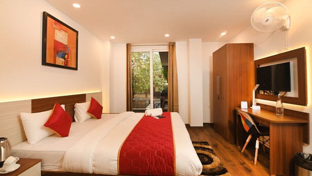 A hotel room with a bed, a desk, a TV, and a window with a view of greenery. - Limewood Stay - Cyber Hub (Gurugram)