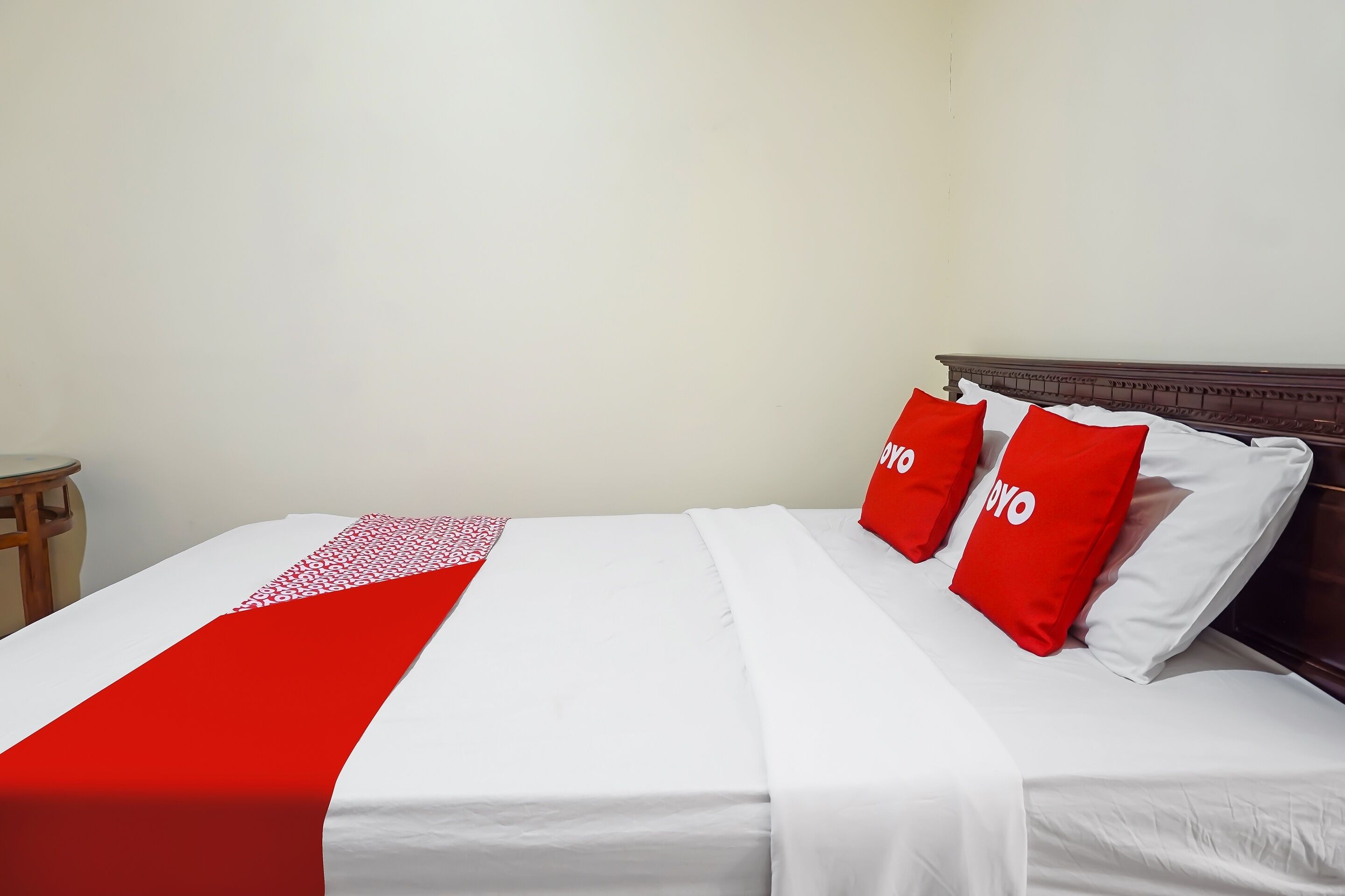 Een opgemaakt bed met wit en rode beddengoed, een houten hoofdbord en een klein houten kruk links.