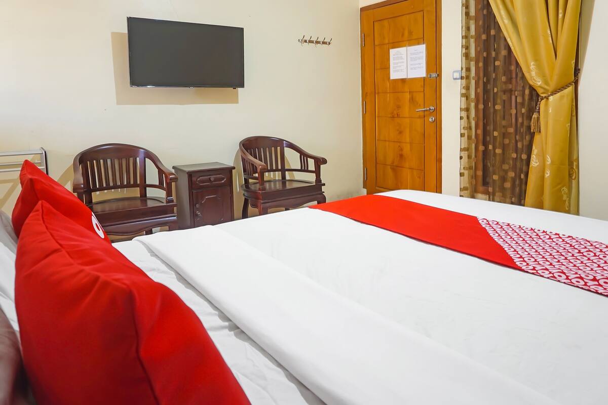 Habitación de hotel con cama, dos sillas de madera, una mesita, un televisor y una puerta con un cartel.