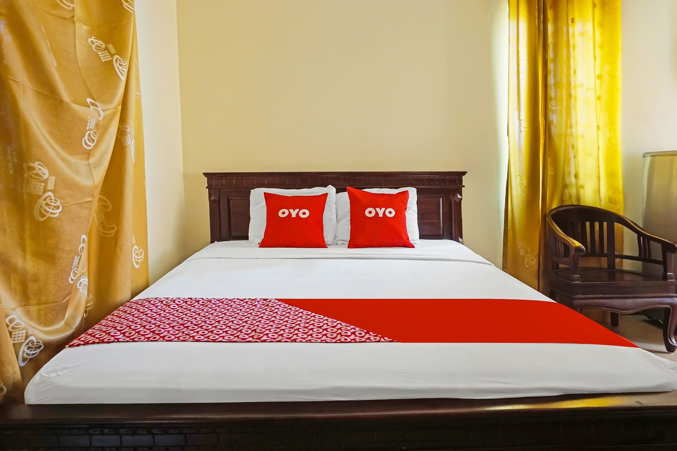 A hotel room with a bed, two red pillows with "OYO" text, a red and white bedspread, and yellow curtains.