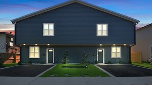 A two-story residential building with a dark blue exterior, two front doors, and a small front yard with grass and shrubs.