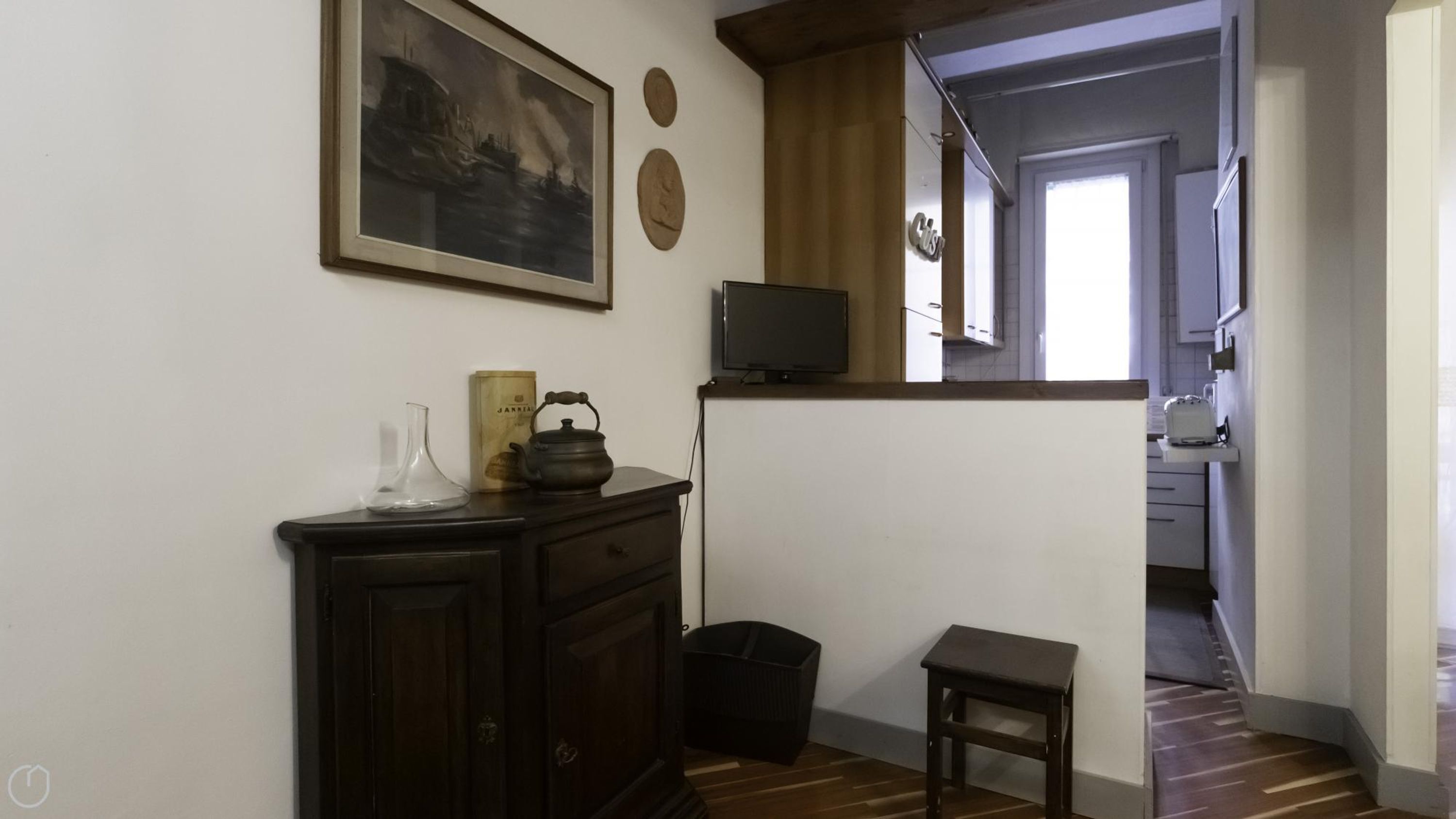 A room with a dark wooden cabinet, a small stool, a black trash bin, a television on a stand, and a framed painting on the wall.
