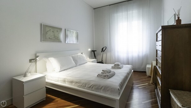 A modern bedroom with a large bed, white nightstands, a wooden dresser, and a large window with sheer curtains. - Dupr 5 (Milan)