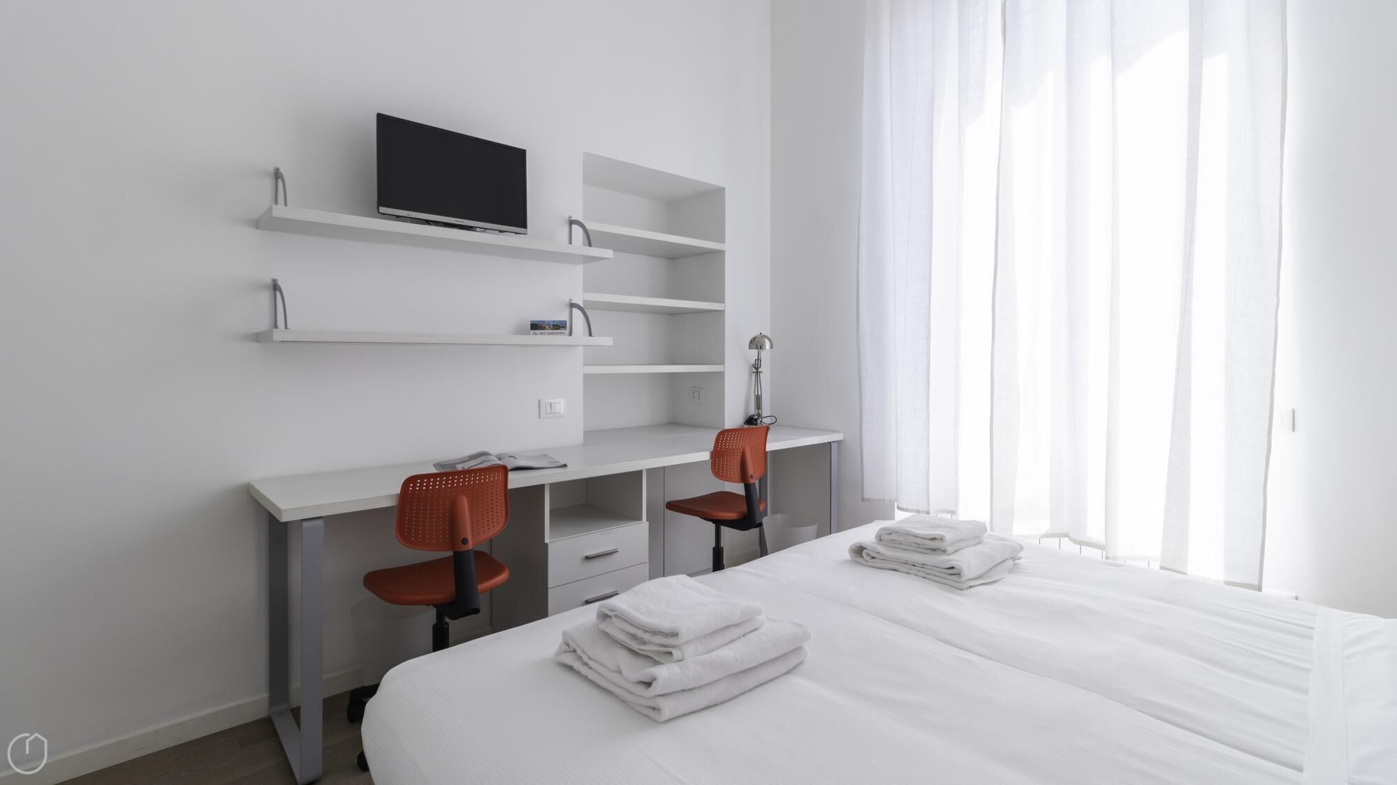A modern hotel room with a bed, two chairs, a desk with a TV, and a window with sheer curtains.