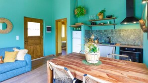 A room with a wooden dining table, a blue sofa, a kitchen with a tiled backsplash, and a hanging plant. - Bibi - Danilia Cottages (Paxos)