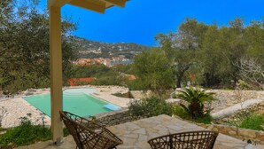 A pool area with two wicker chairs, a stone patio, and a view of a hillside town. - Bibi - Danilia Cottages (Paxos)