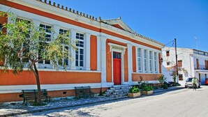 A building with a red door and white columns, a bench, and potted plants on the sidewalk. - Bibi - Danilia Cottages (Paxos)