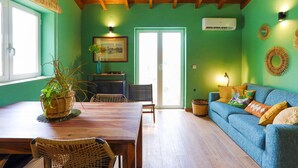 A room with a wooden ceiling, green walls, a blue sofa, a dining table with chairs, a small kitchen area, and a window with blinds. - Sofi - Danilia Cottages (Paxos)
