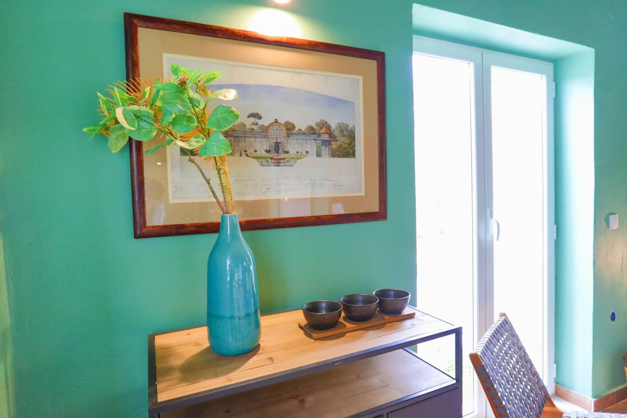 A room with teal walls, a framed landscape painting, a blue vase with a plant, and a wooden table with three bowls.
