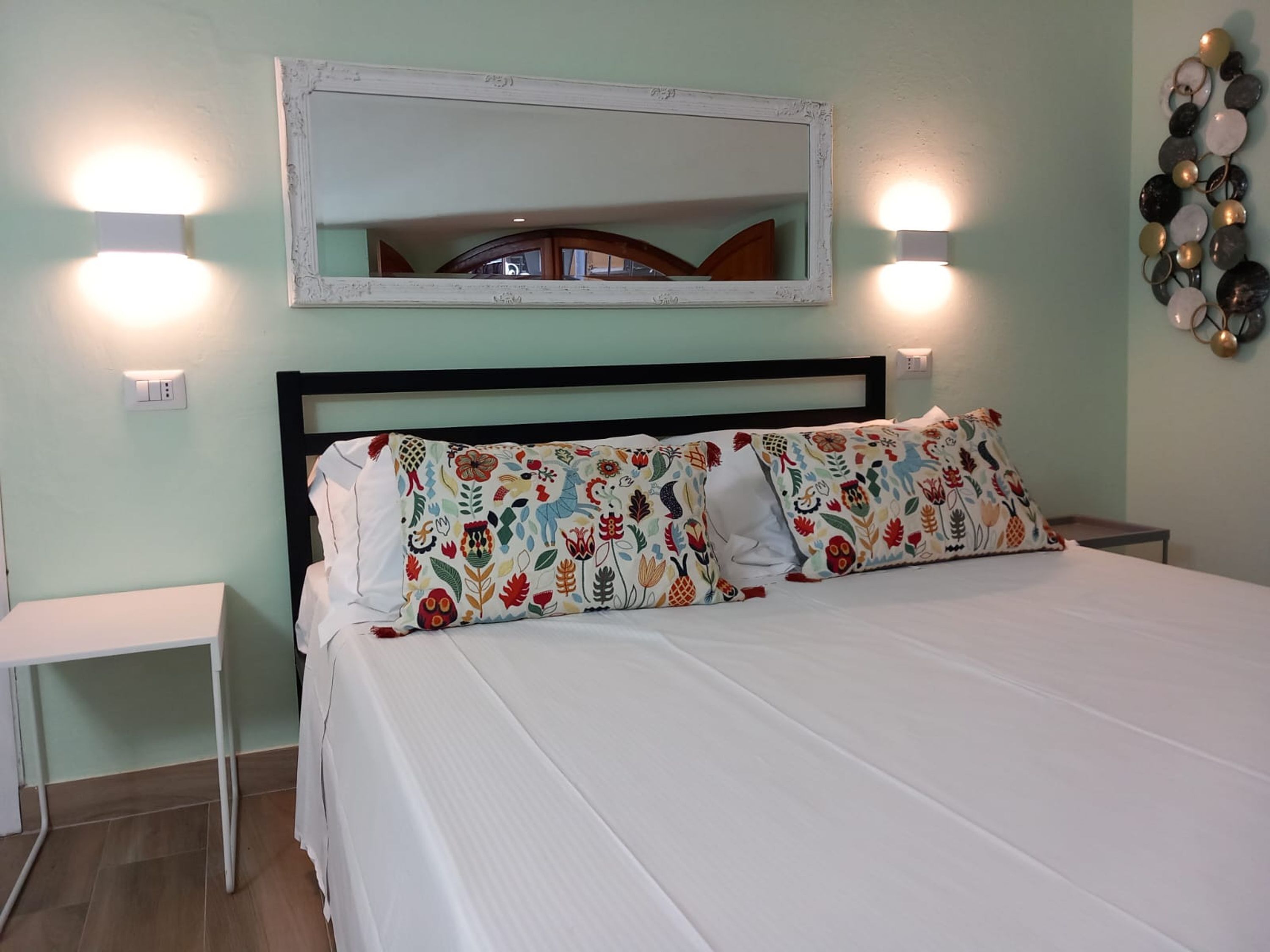 Un lit bien fait, agrémenté d’oreillers à motifs, une table de chevet, un miroir fixé au mur et deux appliques murales.