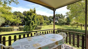 Une terrasse extérieure avec une table recouverte d’une nappe à motifs, entourée de chaises blanches, le tout se fondant dans un décor composé d’un jardin luxuriant et d’une clôture en bois.