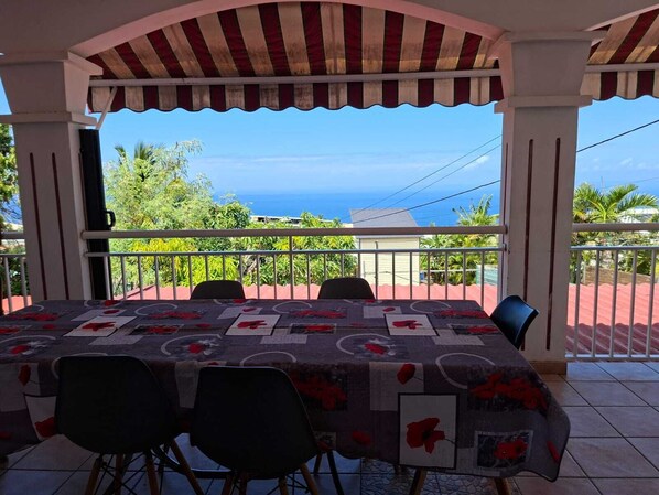 A covered outdoor dining area with a table set for six, overlooking a scenic view of the ocean and greenery.