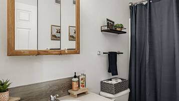 Un baño con un lavamanos blanco, armarios de madera, un espejo con luces, un inodoro, una cortina de ducha oscura y una pequeña estantería con artículos de aseo.