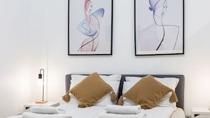 A neatly made bed with white linens and two brown pillows, a bedside lamp, and two abstract art pieces on the wall. - Venus Apartments - King street IV (Budapest)