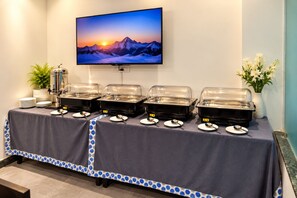 A buffet setup with four covered food dispensers, a television displaying a mountain landscape, and a vase with white flowers on a table. - The Altruist New Town Hotel (Barasat)