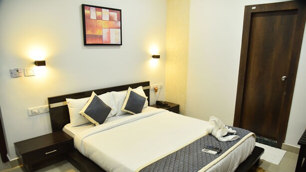 A neatly made bed with pillows, a bedside table, a wall-mounted light, and a door. - The Altruist New Town Hotel (Barasat)