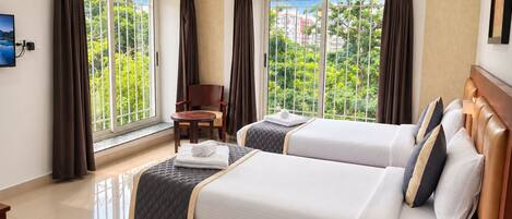A hotel room with two beds, a chair, a table, and large windows with curtains.