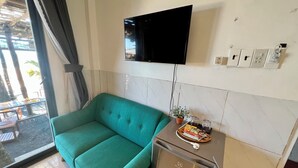 A teal sofa, a small fridge, a TV mounted on the wall, and a window with a view of outdoor seating. - SOHO HOMESTAY PHU QUOC (Phu Quoc)