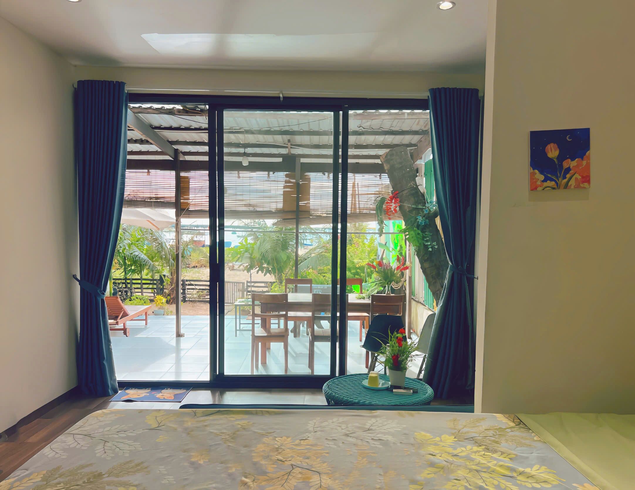 A room with a sliding glass door leading to an outdoor area with a table and chairs, a potted plant, and a view of greenery.