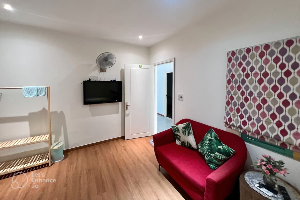 Ein Zimmer mit Holzboden, einem roten Sofa mit grünen Kissen, einem Wandfernseher, einem Standventilator und einer Wand mit Muster.