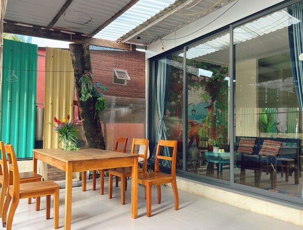 A dining area with a wooden table and chairs, a large window with a view of a street scene, and a tree with hanging plants.