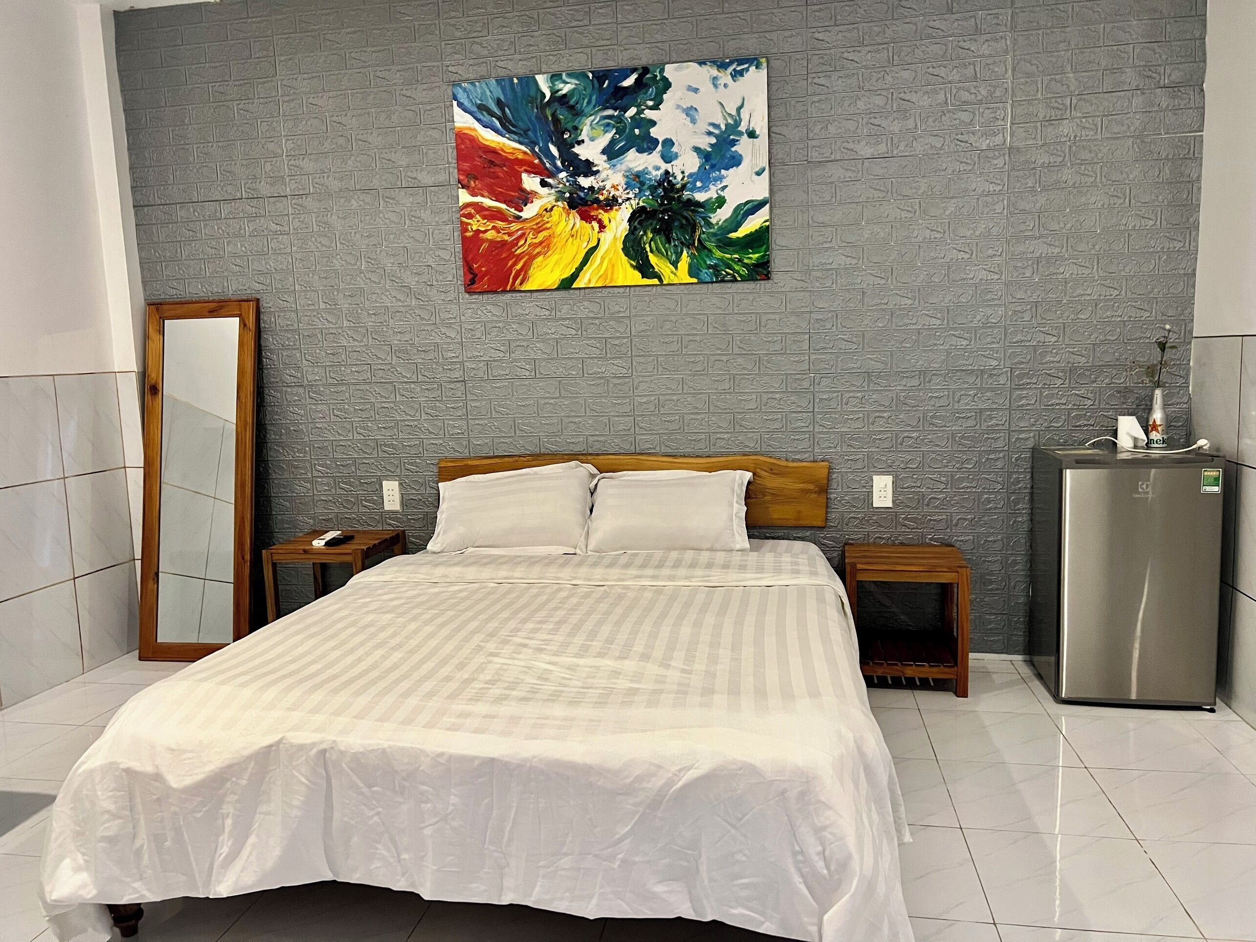 A hotel room with a bed, a nightstand, a mirror, a refrigerator, and a colorful painting on the wall.