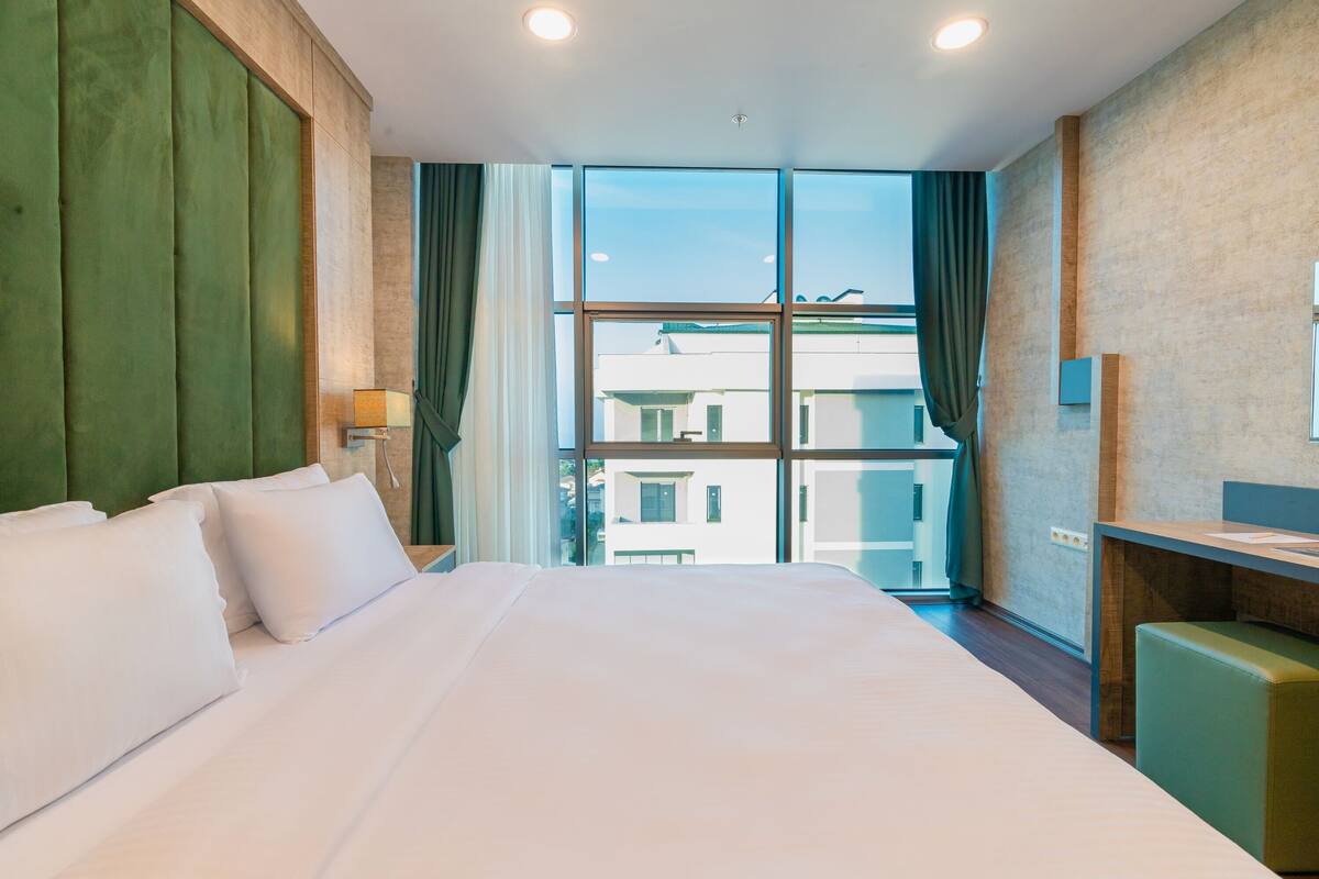 A hotel room with a large bed, a desk, and a view of buildings through the window.