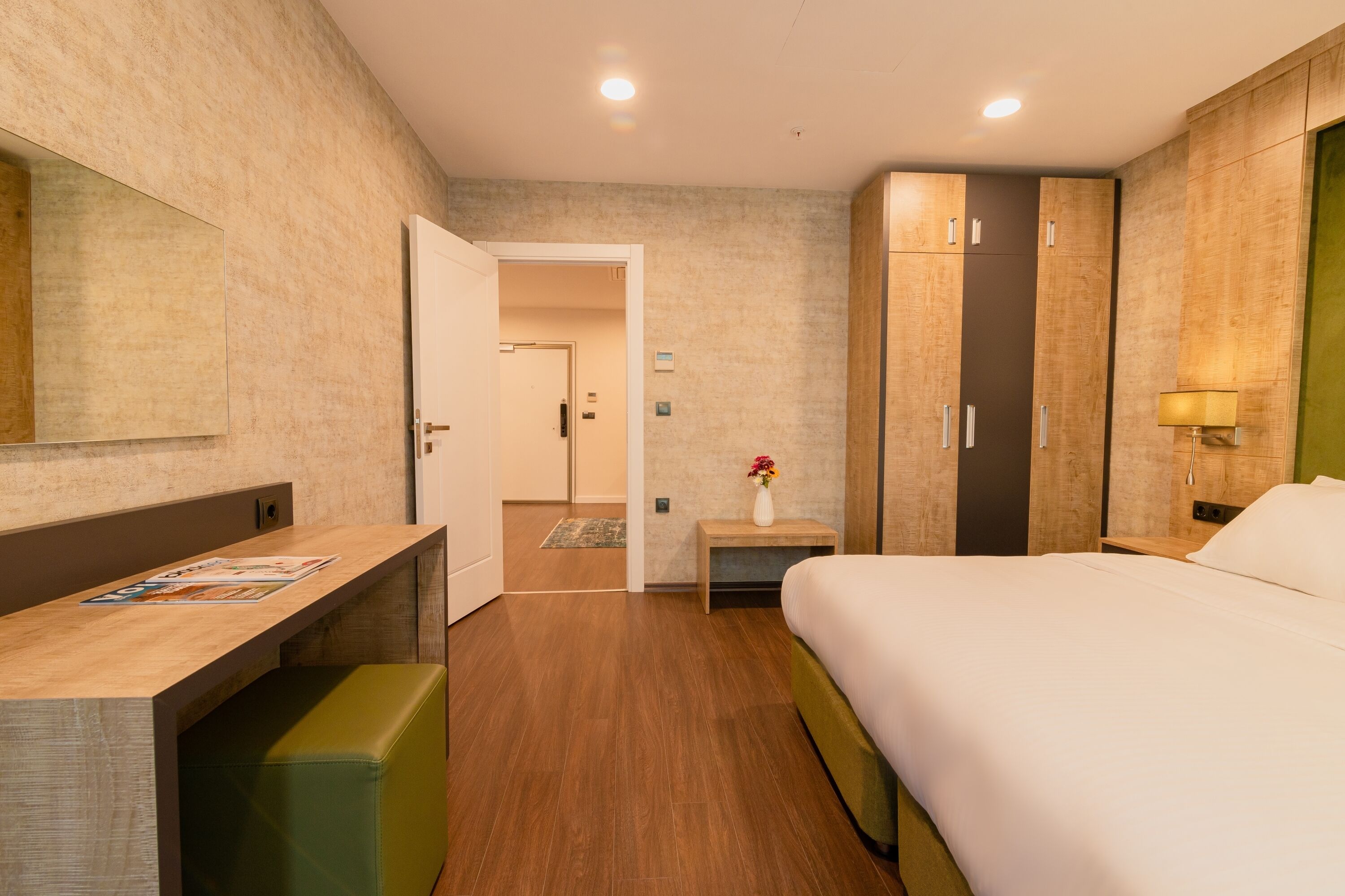 Habitación de hotel con un escritorio de madera, un banco verde, una cama, un armario y un espejo en la pared.