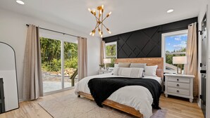 A modern bedroom with a large bed, a nightstand, a dresser, and a chandelier. - 4 King Luxury MTN Home | 3 min Downtown Black MTN | 17 min to Asheville (Black Mountain)