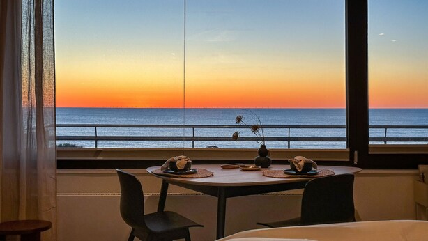 A room with a view of the ocean at sunset, a dining table with plates and a vase, and two beds. - Apartment Ocean Suite (Wenningstedt)