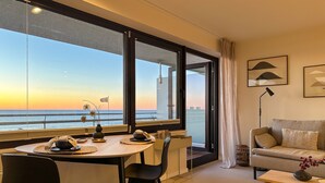 A modern living room with a dining area, large windows with a view of the sea, a sofa, and a lamp. - Apartment Ocean Suite (Wenningstedt)