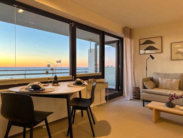 A modern living room with a dining area, large windows with a view of the sea, a sofa, and a lamp.
