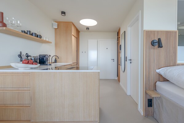 Une cuisine moderne avec des meubles en bois, un plan de travail blanc et un four encastré. On y trouve un petit coin repas avec une table et des chaises blanches, ainsi qu’un banc en bois avec un coussin.