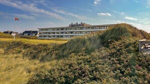 A modern multi-story building with a flat roof, surrounded by grassy terrain and dense shrubbery. - Apartment Ocean Suite (Wenningstedt)