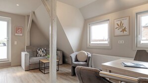 A modern loft-style room with a sloped ceiling, a grey sofa, a small table, a grey armchair, and a wooden dining table. - Appartement Hummer (Westerland)