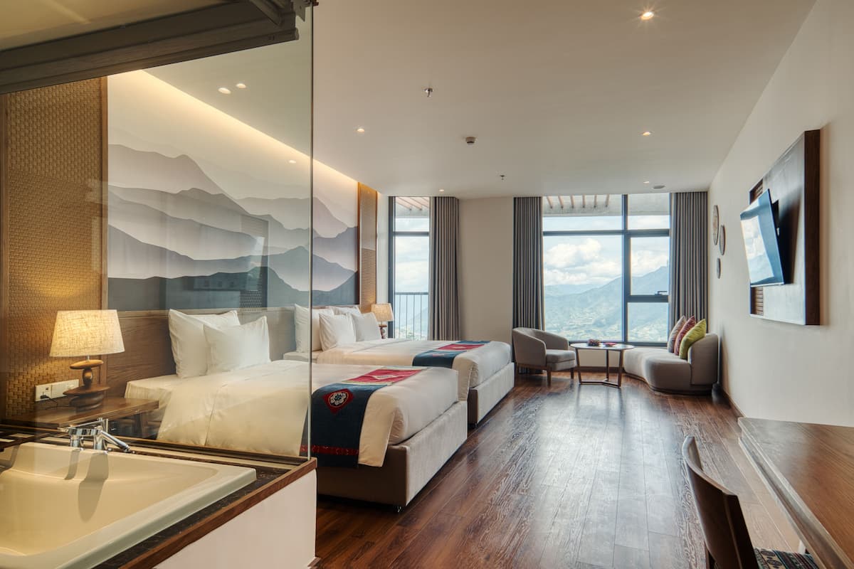 Ein Hotelzimmer mit zwei Betten, einer Sitzecke inklusive Sofa und Sesseln, einem großen Fenster mit Blick auf die Berge und einem an der Wand befestigten Fernseher.