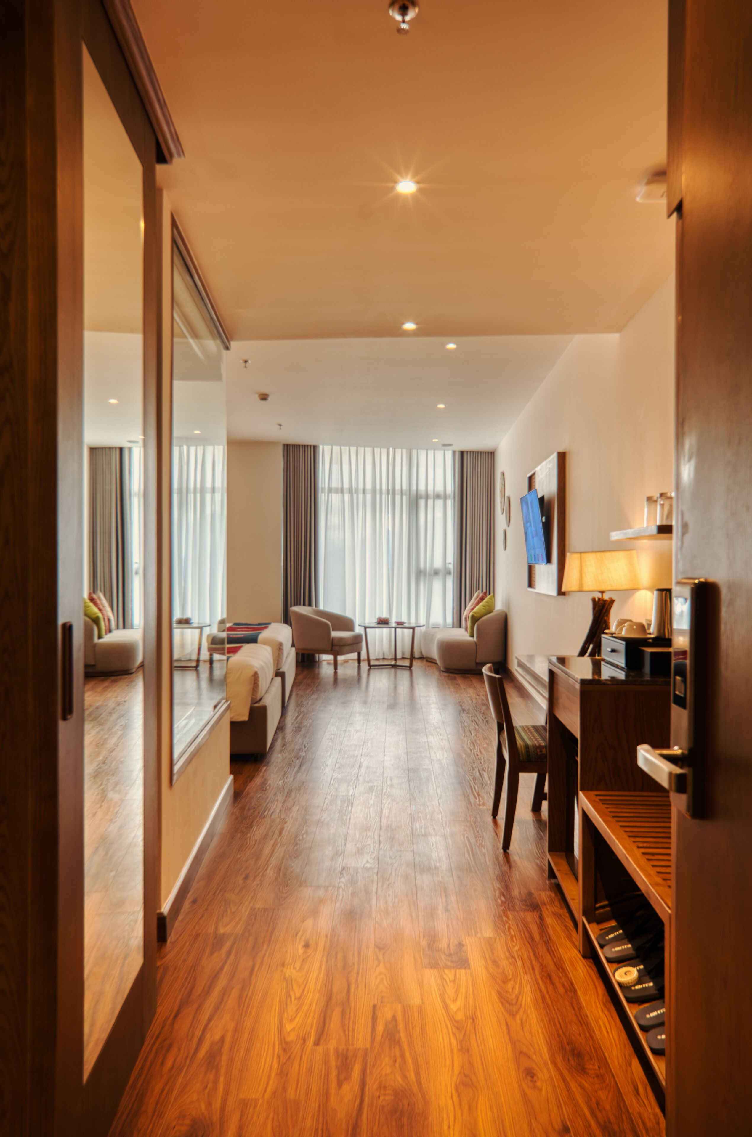 Hotelkamer met houten vloer, bureau met lamp, stoel, bank en televisie aan de muur.