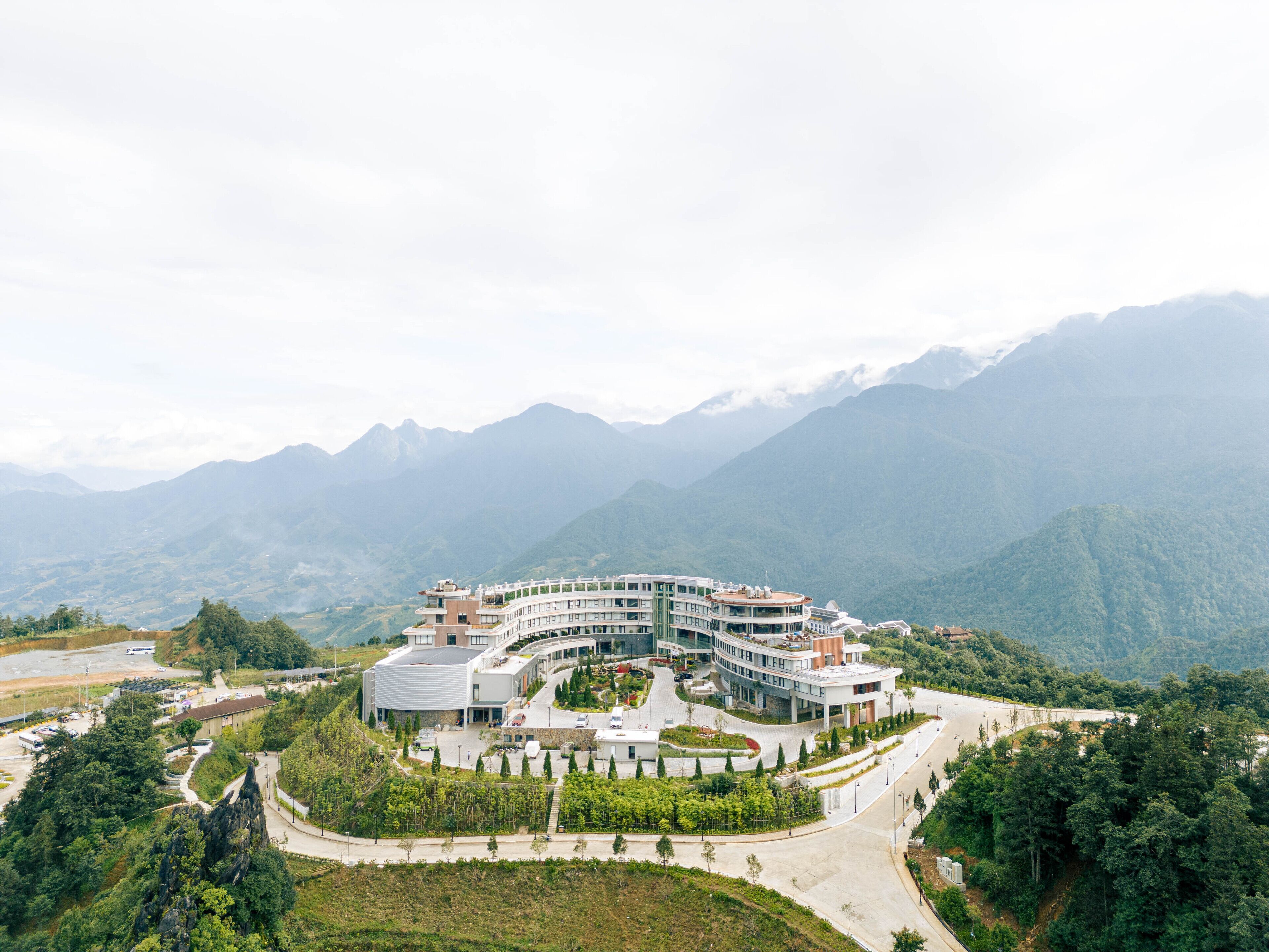 A modern hotel with a curved structure, surrounded by lush greenery and mountains in the background.
