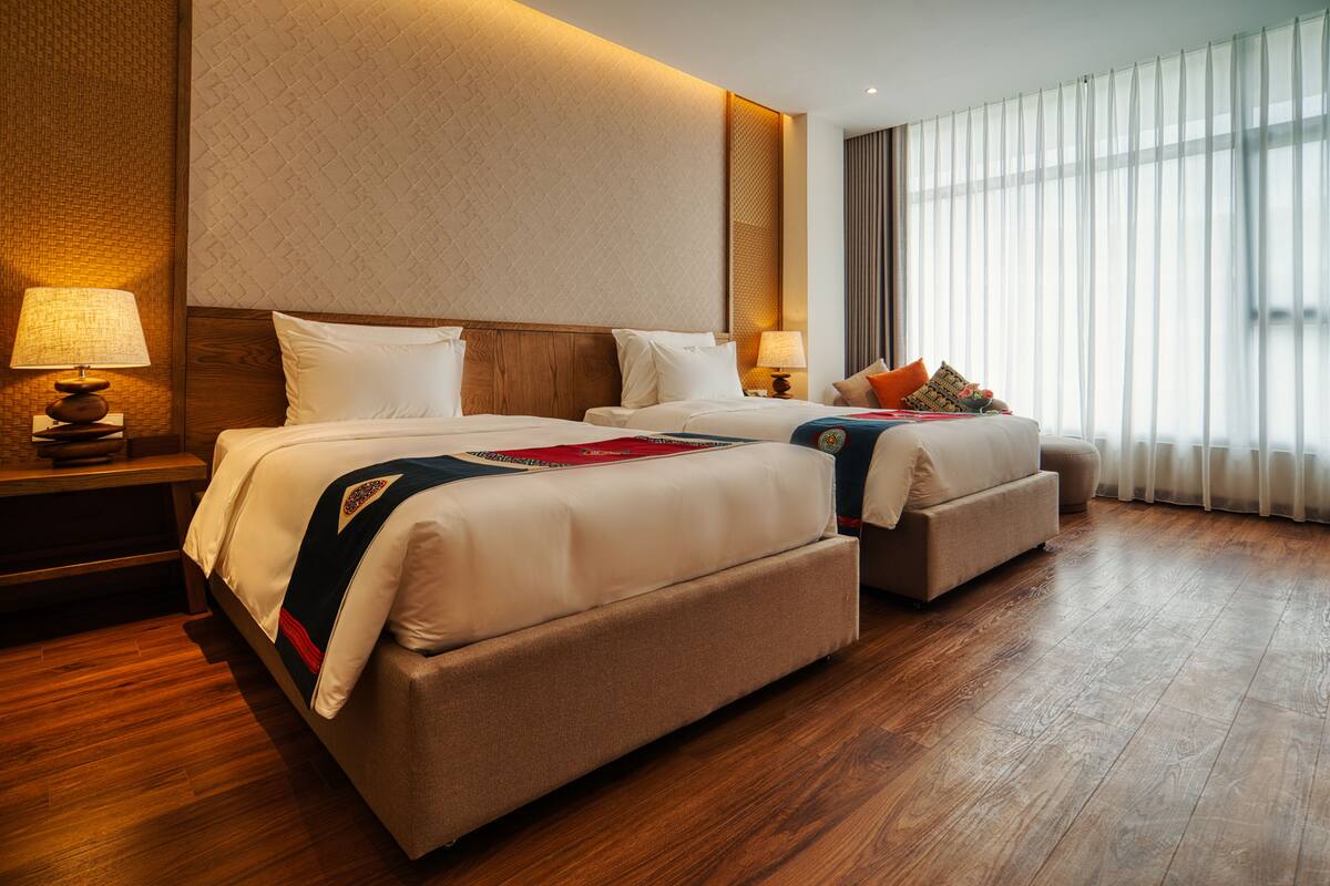 Ein Hotelzimmer mit zwei Betten, Holzboden und einem großen Fenster mit Vorhängen.