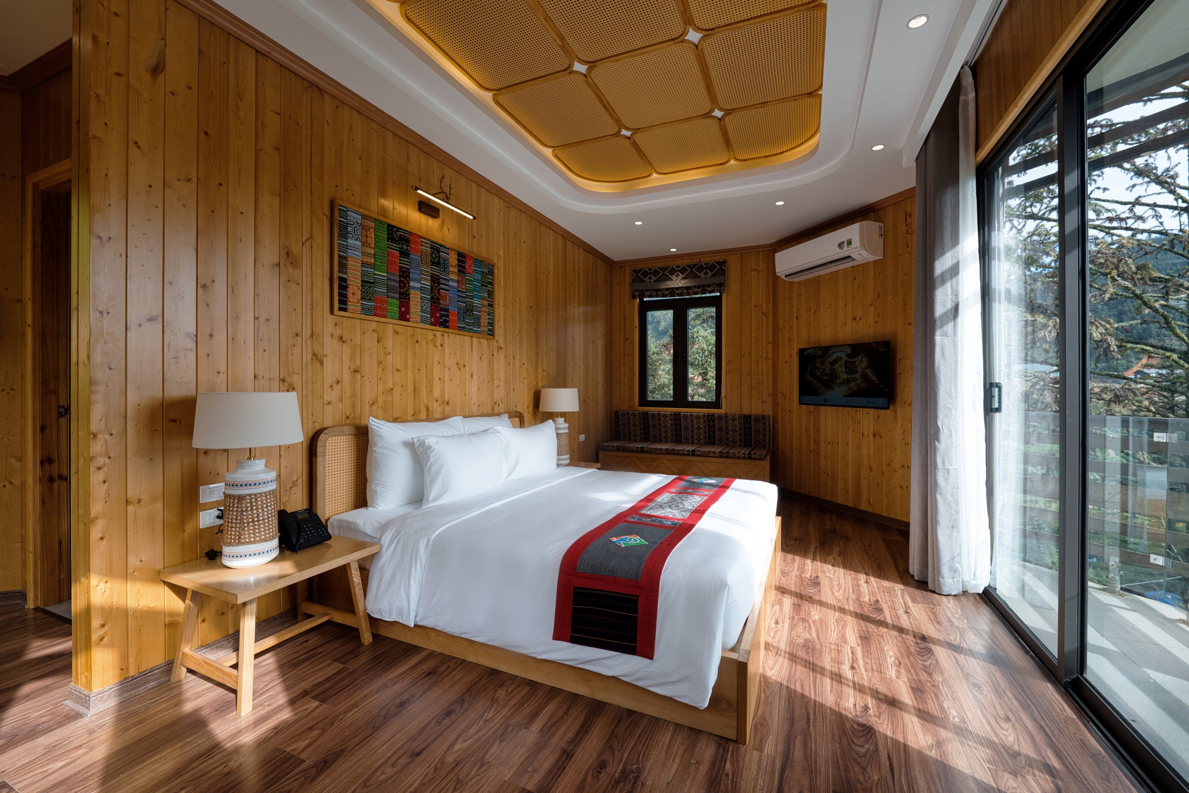 A wooden-paneled bedroom with a large bed, a bedside table with a lamp, a wall-mounted air conditioner, and a large window with a view of trees.