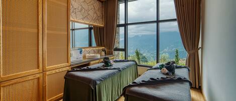 Un cuarto de masajes con dos camillas, una pared de madera con diseño trenzado y un ventanal con vista a la montaña.