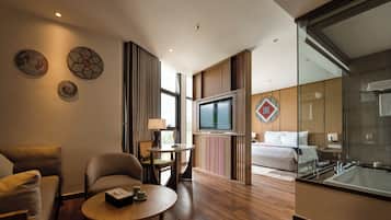 A modern hotel room with a sofa, armchair, small table, TV, and a large window.