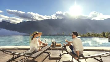 A couple relaxing by a pool with a mountain view, a table with drinks and a bucket, and lounge chairs.