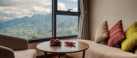 Una habitación con sofá, una mesita redonda con frutas y una servilleta, y ventana con vistas a la montaña.