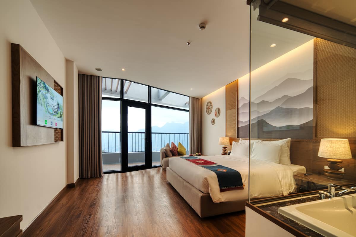 Ein modernes Hotelzimmer mit einem großen Bett, einem Badezimmer mit Badewanne, einem großen Fenster mit Blick und einem an der Wand befestigten Kunstwerk.