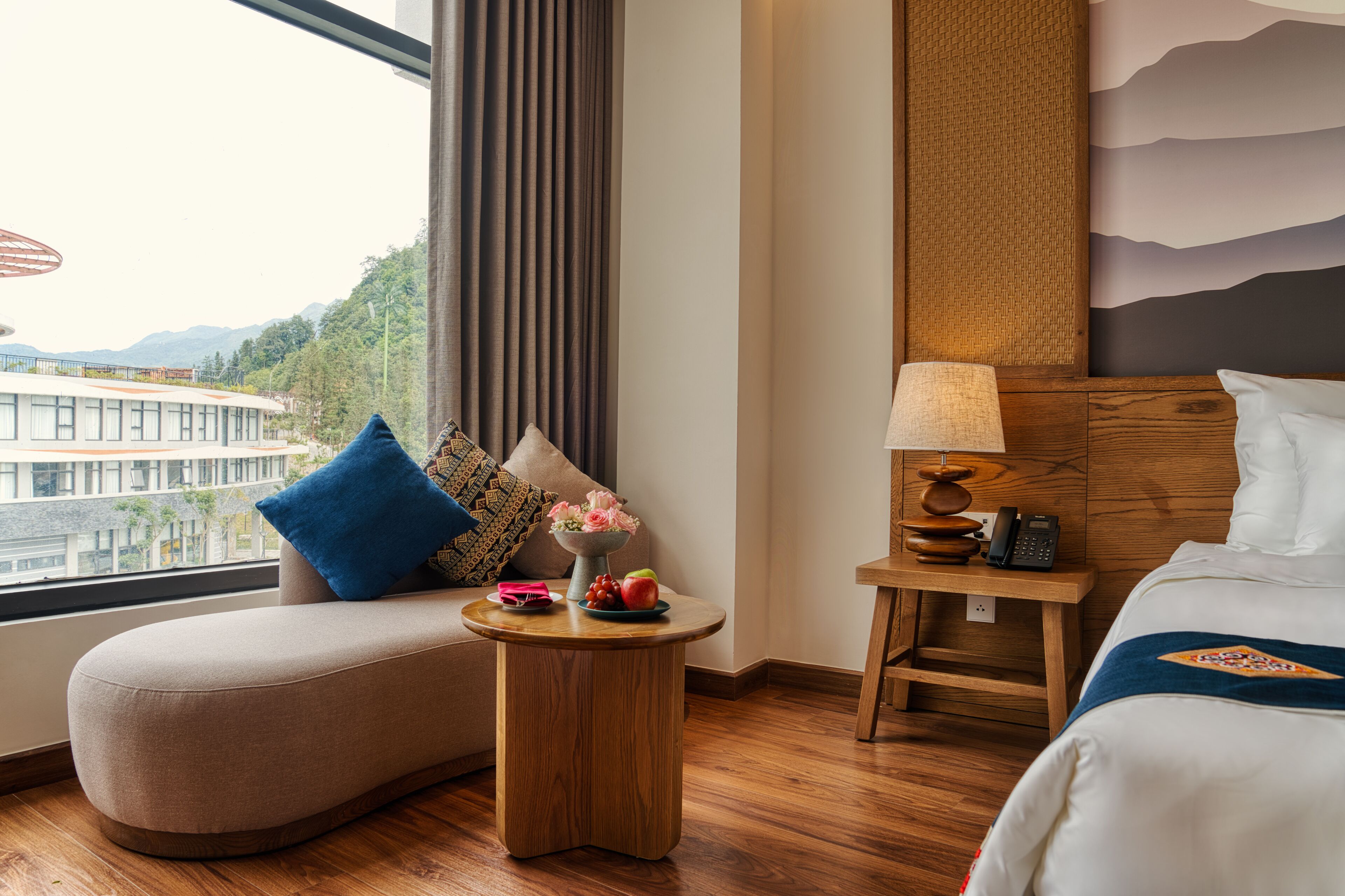 A hotel room with a sofa, a small round table with a lamp, a bedside table with a phone, and a bed with a headboard.
