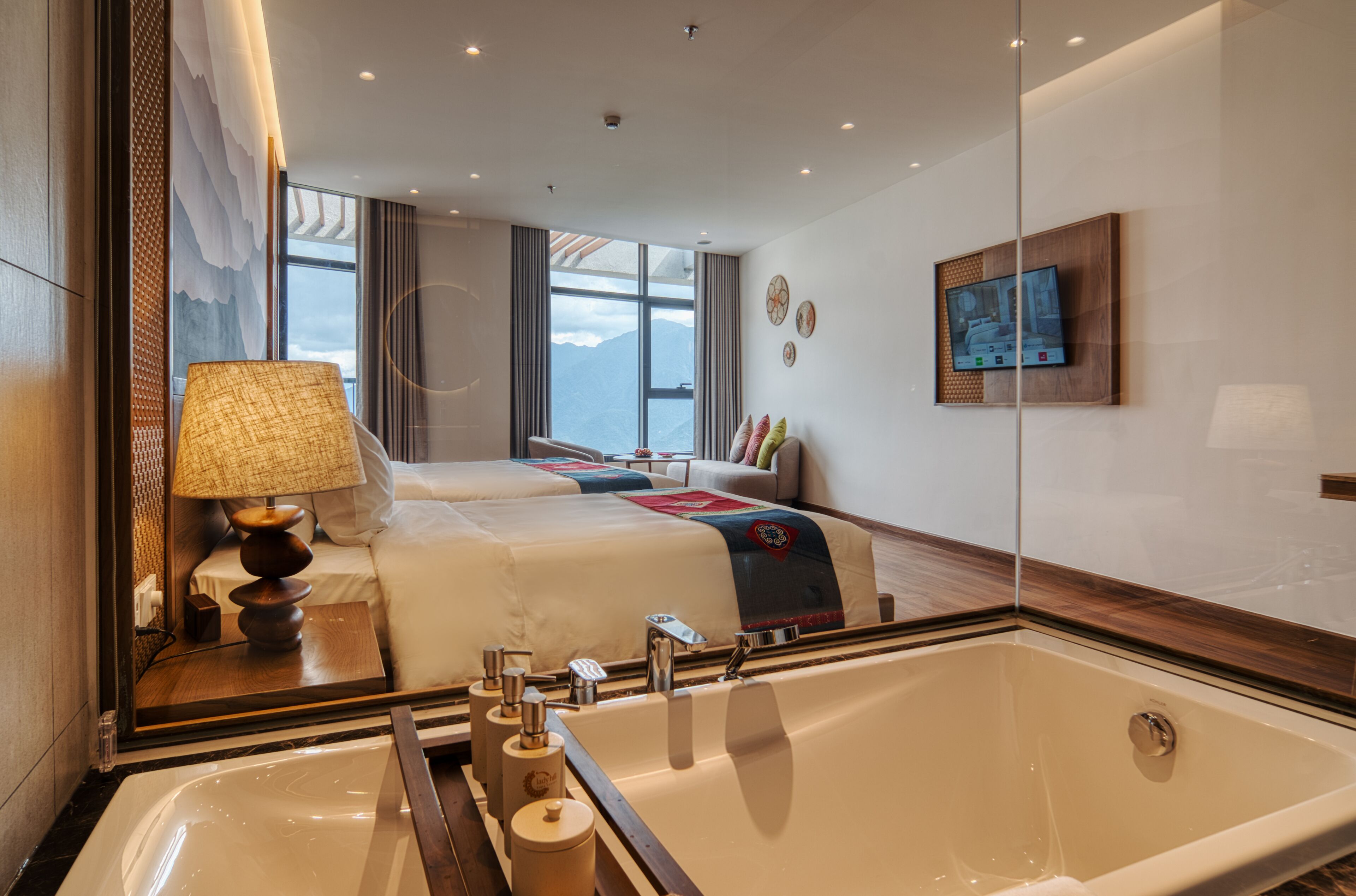 A hotel room with a large bathtub, two beds, a TV on the wall, and a view of mountains through the window.